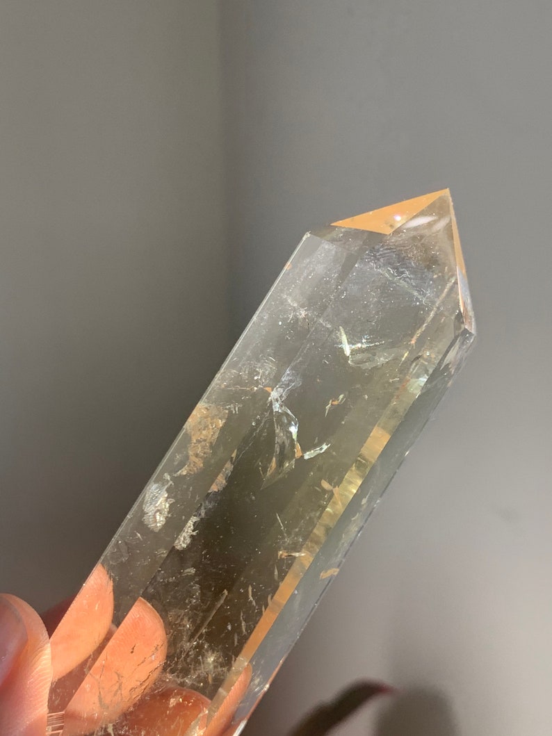 May include: A clear quartz crystal point with a golden hue. The crystal is long and slender with a pointed tip. It has a natural, raw look.