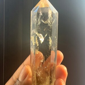 May include: A large, clear quartz crystal point with a golden hue. The crystal is faceted and has a pointed tip. The crystal is being held in a hand.