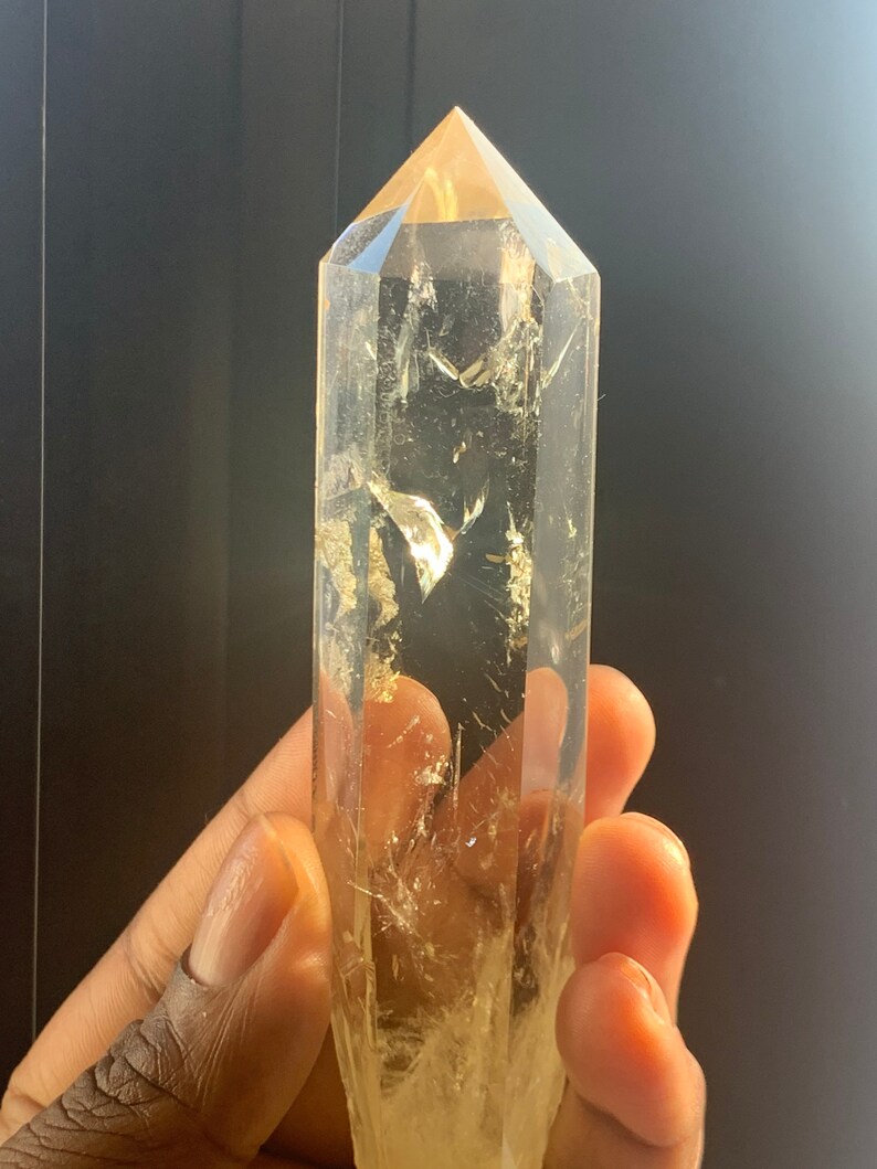 May include: A large, clear quartz crystal point with a golden hue. The crystal is faceted and has a pointed tip. The crystal is being held in a hand.