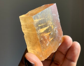 Pode incluir: Um grande cristal de calcita amarelo claro com forma retangular. O cristal tem uma ligeira tonalidade laranja e é translúcido.
