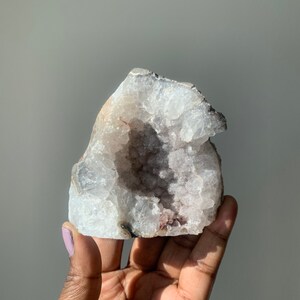 K&ouml;nnte beinhalten: Ein wei&szlig;er Kristallgeode mit einer gro&szlig;en &Ouml;ffnung, die einen Cluster kleinerer Kristalle im Inneren freilegt. Der Geode hat eine raue, nat&uuml;rliche Textur und ist hellviolett.