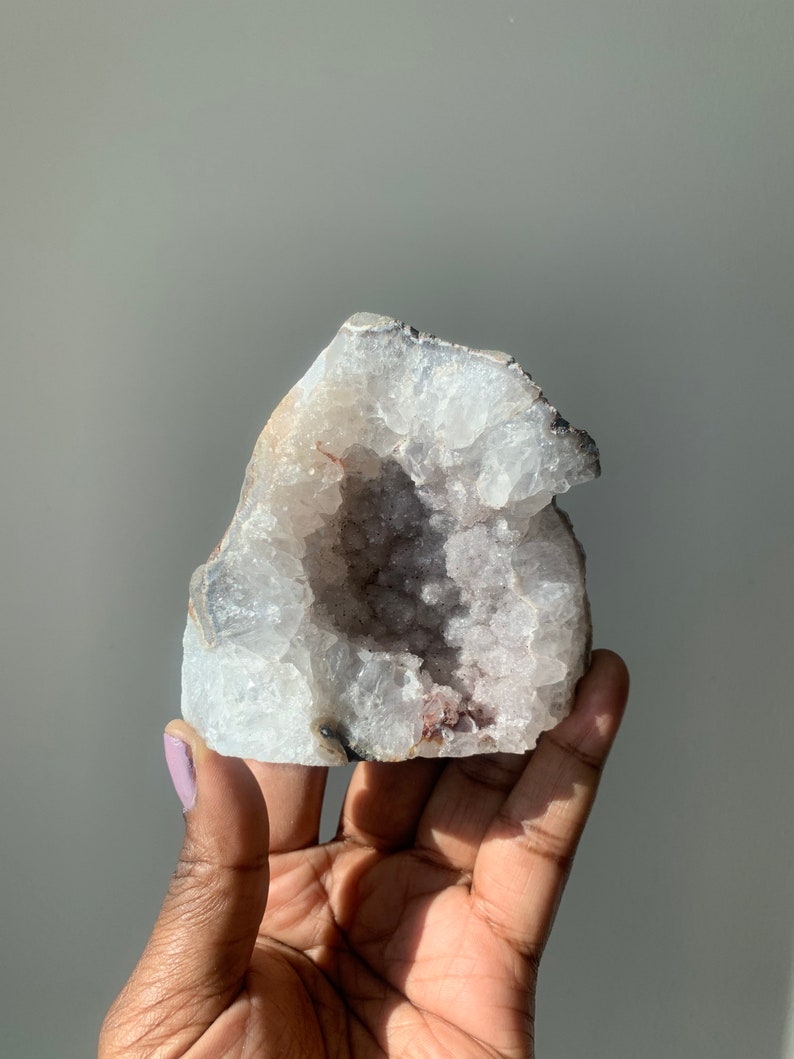 K&ouml;nnte beinhalten: Ein wei&szlig;er Kristallgeode mit einer gro&szlig;en &Ouml;ffnung, die einen Cluster kleinerer Kristalle im Inneren freilegt. Der Geode hat eine raue, nat&uuml;rliche Textur und ist hellviolett.