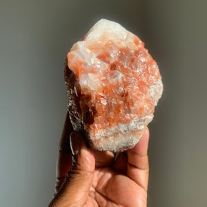 May include: A raw aragonite crystal cluster, showcasing a mix of white, orange, and reddish-brown hues. The natural stone has a rough, textured surface and is held in a hand, with a neutral background.