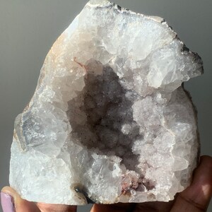 K&ouml;nnte beinhalten: Ein gro&szlig;er, wei&szlig;er Geodenkristall mit einem hohlen Zentrum, das mit kleinen, violetten Kristallen ausgekleidet ist. Die Geode ist teilweise ge&ouml;ffnet und zeigt die Kristalle im Inneren.