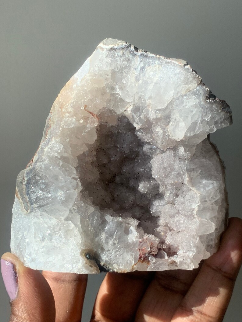 K&ouml;nnte beinhalten: Ein gro&szlig;er, wei&szlig;er Geodenkristall mit einem hohlen Zentrum, das mit kleinen, violetten Kristallen ausgekleidet ist. Die Geode ist teilweise ge&ouml;ffnet und zeigt die Kristalle im Inneren.