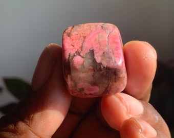 May include: A pink and gray tumbled stone with a smooth, polished surface. The stone has a unique pattern of pink and gray veins.