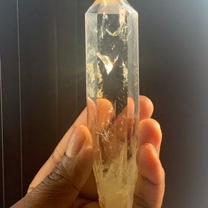 May include: A clear quartz crystal point with a golden hue. The crystal is long and slender, with a pointed tip and a flat base. The crystal is held in a hand, and the light shines through it, creating a beautiful sparkle.