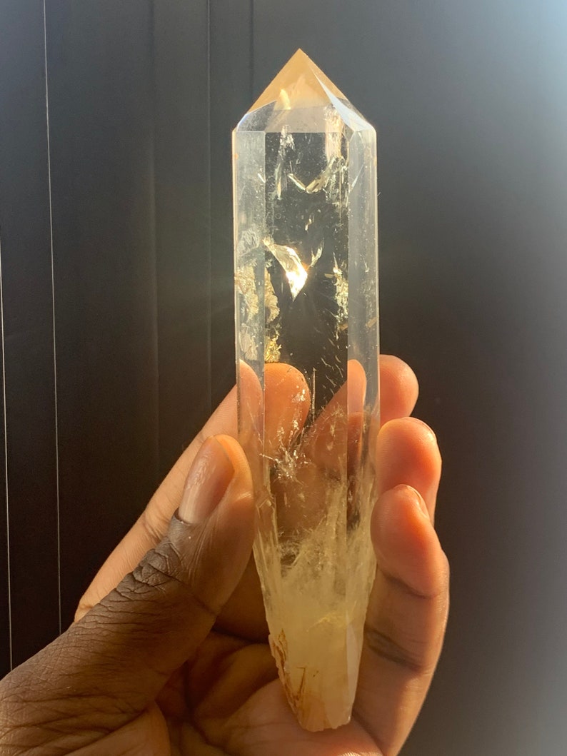 May include: A clear quartz crystal point with a golden hue. The crystal is long and slender, with a pointed tip and a flat base. The crystal is held in a hand, and the light shines through it, creating a beautiful sparkle.