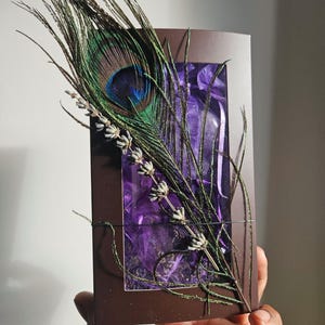 May include: A brown box with a clear window, showcasing a purple fabric with dried lavender flowers and a peacock feather. The box is held in a hand.