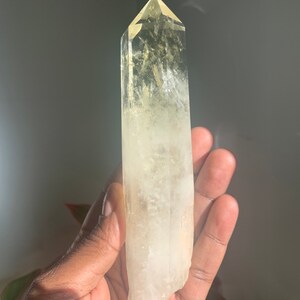 Pode incluir: Um cristal claro e alongado com uma ponta. O cristal transita de um amarelo p&aacute;lido no topo para um branco leitoso na base. O cristal &eacute; segurado em uma m&atilde;o, com um fundo neutro.
