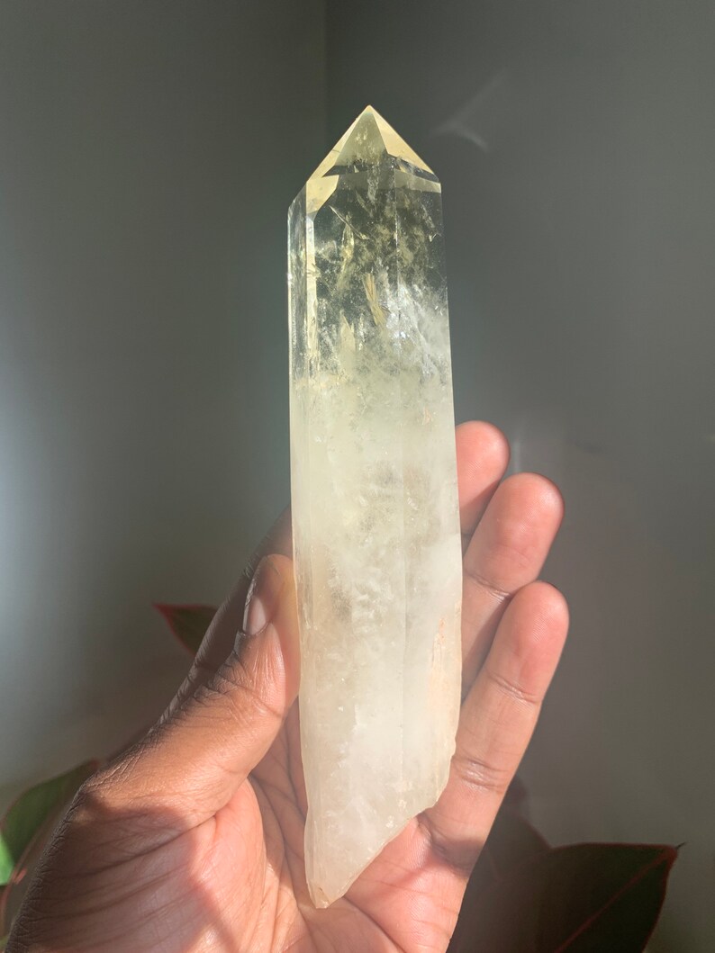 Pode incluir: Um cristal claro e alongado com uma ponta. O cristal transita de um amarelo p&aacute;lido no topo para um branco leitoso na base. O cristal &eacute; segurado em uma m&atilde;o, com um fundo neutro.