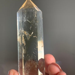 May include: A clear quartz crystal point with a golden hue. The crystal is faceted and has a pointed tip. The crystal is held in a hand.