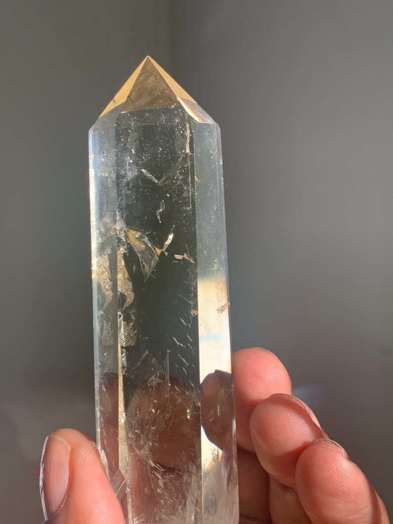 May include: A clear quartz crystal point with a golden hue. The crystal is faceted and has a pointed tip. The crystal is held in a hand.