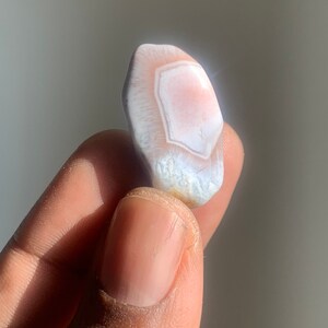 以下が含まれることがあります： 不規則な形をした、白とピンクの小さなアゲートストーン。層状のパターンがあります。石は手で持たれています。
