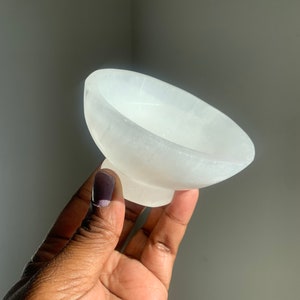 May include: A small, white selenite bowl with a smooth, polished surface. The bowl is slightly curved and has a rounded base.