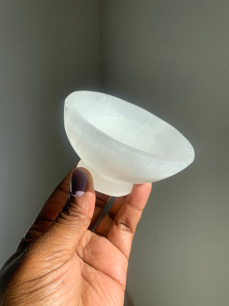 May include: A small, white selenite bowl with a smooth, polished surface. The bowl is slightly curved and has a rounded base.