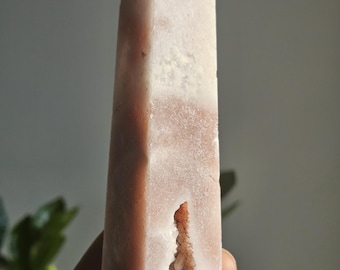 May include: A pink quartz crystal obelisk with a pointed top. The crystal has a rectangular shape with a hollowed-out section on the side. The obelisk is a light pink color with white and darker pink variations.