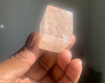 Pode incluir: Um cristal translúcido, rosa claro, em forma de cubo com um brilho ligeiramente iridescente.