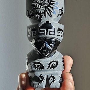 May include: A small black obsidian sculpture of a person with intricate carvings. The sculpture is wearing a headdress with a stylized bird design and has geometric patterns on the body.
