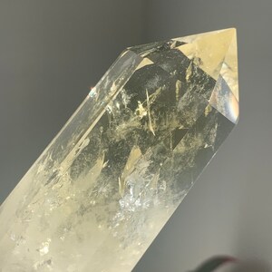 Pode incluir: Close-up de uma ponta de cristal de quartzo transparente com uma ligeira tonalidade amarela. O cristal &eacute; facetado e tem inclus&otilde;es internas, com a luz refletindo nas superf&iacute;cies. O fundo &eacute; cinza desfocado.