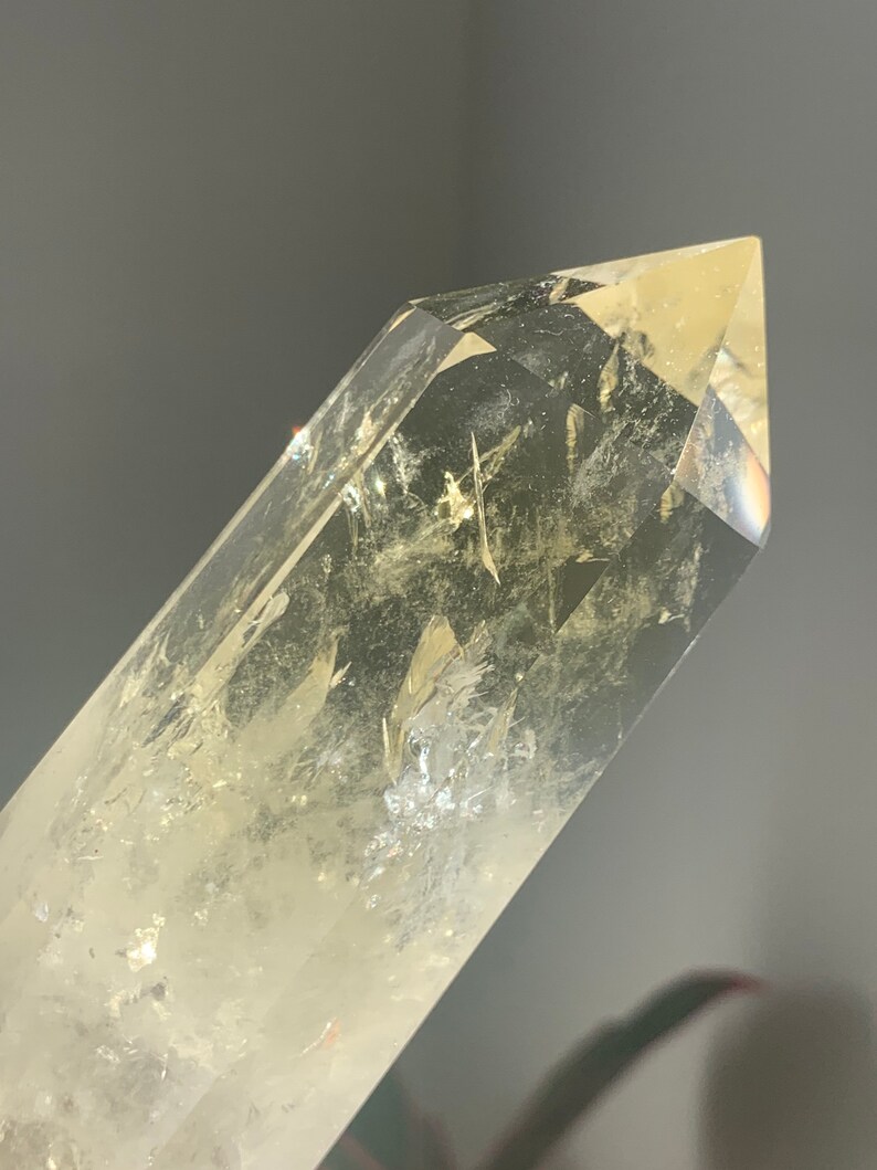 Pode incluir: Close-up de uma ponta de cristal de quartzo transparente com uma ligeira tonalidade amarela. O cristal &eacute; facetado e tem inclus&otilde;es internas, com a luz refletindo nas superf&iacute;cies. O fundo &eacute; cinza desfocado.
