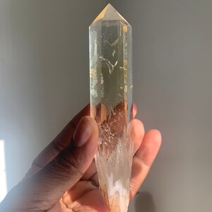 May include: A clear quartz crystal point with a golden hue. The crystal is pointed at the top and has a natural, rough texture. The crystal is approximately 6 inches long.