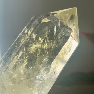 Pode incluir: Uma ponta de cristal de quartzo transparente com um tom amarelo. O cristal tem uma ponta afiada e facetada e inclus&otilde;es internas. A luz refrata-se atrav&eacute;s do cristal, criando um efeito brilhante. Esp&eacute;cime geol&oacute;gico natural.
