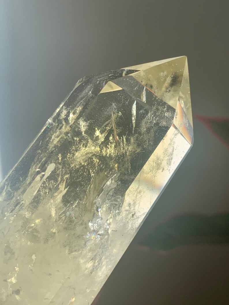 Pode incluir: Uma ponta de cristal de quartzo transparente com um tom amarelo. O cristal tem uma ponta afiada e facetada e inclus&otilde;es internas. A luz refrata-se atrav&eacute;s do cristal, criando um efeito brilhante. Esp&eacute;cime geol&oacute;gico natural.