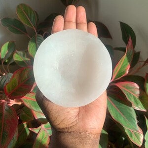 May include: A white selenite bowl, also known as a sound bowl, is held in a person's hand. The bowl is smooth and has a slightly rounded shape.