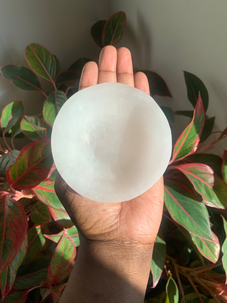 May include: A white selenite bowl, also known as a sound bowl, is held in a person's hand. The bowl is smooth and has a slightly rounded shape.