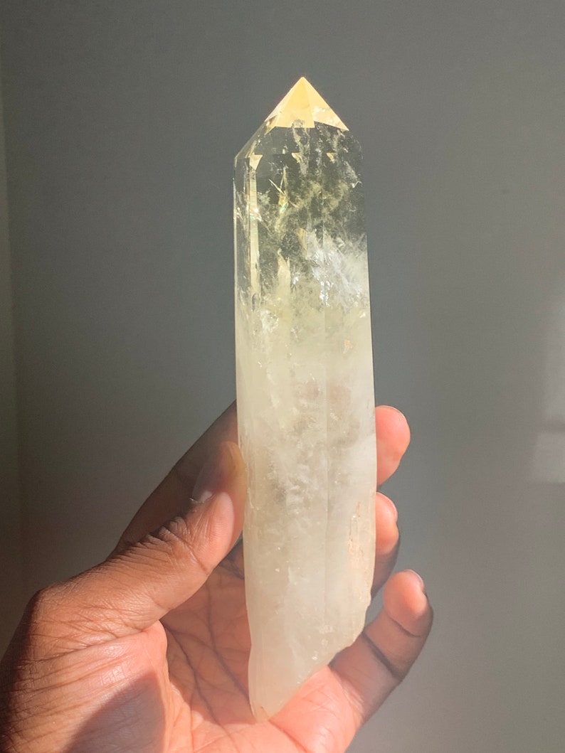 Pode incluir: Uma ponta de cristal de quartzo transparente com uma ponta amarela. O cristal &eacute; segurado em uma m&atilde;o e &eacute; parcialmente transl&uacute;cido, permitindo que a luz passe por ele. O cristal &eacute; de cor amarelo p&aacute;lido e tem uma apar&ecirc;ncia natural e n&atilde;o polida.