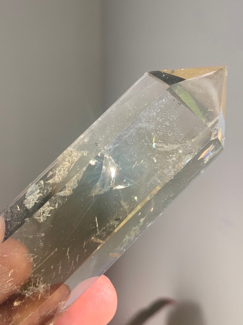 May include: A clear quartz crystal point with a smoky brown hue. The crystal is faceted and has a pointed tip. The crystal is approximately 5 inches long.