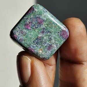 May include: A green and pink gemstone with a square shape. The gemstone has a rough texture and is speckled with small, dark red and blue inclusions.