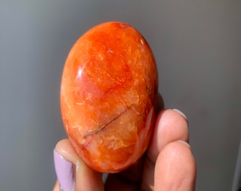 Puede incluir: Una piedra de cornalina lisa y pulida, de color rojo-naranja, con una forma natural y orgánica. La piedra es translúcida y muestra intrincados patrones de vetas y estrías.