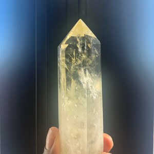 Pode incluir: Uma ponta de cristal de quartzo amarelo claro com um design facetado. O cristal &eacute; segurado em uma m&atilde;o e tem um topo pontiagudo. O cristal &eacute; transl&uacute;cido com algumas inclus&otilde;es internas.