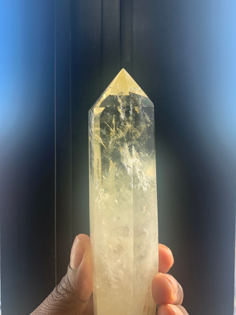 Pode incluir: Uma ponta de cristal de quartzo amarelo claro com um design facetado. O cristal &eacute; segurado em uma m&atilde;o e tem um topo pontiagudo. O cristal &eacute; transl&uacute;cido com algumas inclus&otilde;es internas.