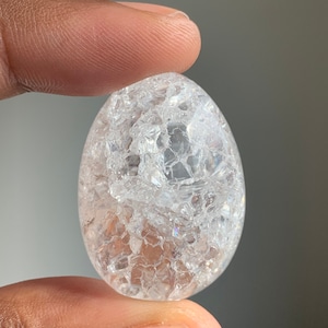 Puede incluir: Un huevo de cristal de cuarzo transparente con una superficie única, agrietada y texturizada. El cristal es translúcido y tiene un ligero brillo iridiscente.