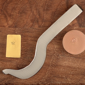 May include: A silver metal stamping tool with a curved handle, a gold metal stamp with a crown design, and a brown leather stamp with a crown design.