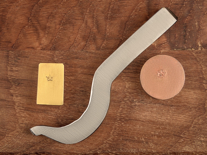 May include: A silver metal stamping tool with a curved handle, a gold metal stamp with a crown design, and a brown leather stamp with a crown design.