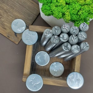 Puede incluir: Una caja de madera llena de cabezales de sellos met&aacute;licos con varios dise&ntilde;os florales. La caja est&aacute; abierta y los cabezales de los sellos est&aacute;n dispuestos en un patr&oacute;n circular. Los cabezales de los sellos son de color plateado y tienen un acabado pulido.