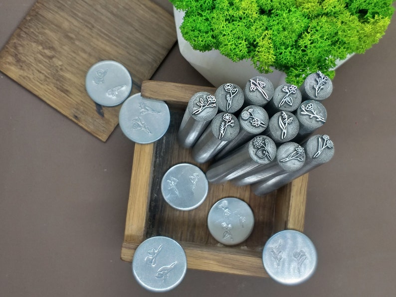 Puede incluir: Una caja de madera llena de cabezales de sellos met&aacute;licos con varios dise&ntilde;os florales. La caja est&aacute; abierta y los cabezales de los sellos est&aacute;n dispuestos en un patr&oacute;n circular. Los cabezales de los sellos son de color plateado y tienen un acabado pulido.