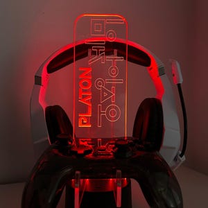 May include: A gaming controller stand with a headset holder. The stand is black with red LED lighting. A clear acrylic panel with text is illuminated in red. The text reads "PLATON" and other characters. The base of the stand has the text "DESIGN 6".