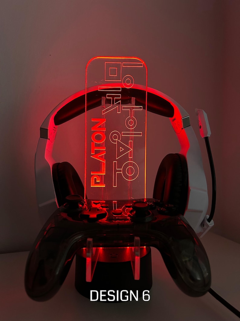 May include: A gaming controller stand with a headset holder. The stand is black with red LED lighting. A clear acrylic panel with text is illuminated in red. The text reads "PLATON" and other characters. The base of the stand has the text "DESIGN 6".