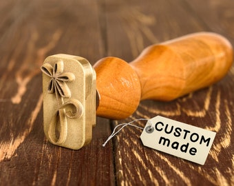 May include: A brass stamp with a floral design and a wooden handle. The stamp is attached to a white tag with the text "CUSTOM made".