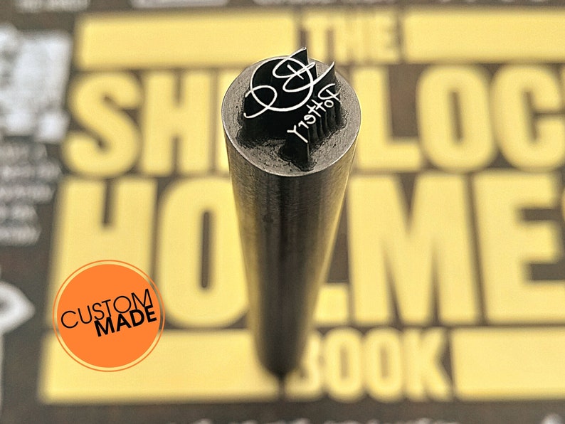 May include: A close-up of a custom-made metal stamp with a black and white design. The stamp is shaped like a cylinder and has a rounded top. The design on the stamp is a stylized letter "B" with the word "Bristol" written below it. The stamp is sitting on a yellow and black book with the words "The Sherlock Holmes Book" printed on it.