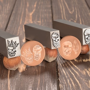 Pode incluir: Quatro carimbos de metal com diferentes desenhos de caveiras. Os carimbos estão sobre uma superfície de madeira com avelãs. Os carimbos são usados para criar impressões em metal.