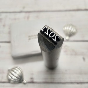May include: A close-up of a metal stamp with the year 2021 engraved on it. The stamp is silver and has a rounded top.