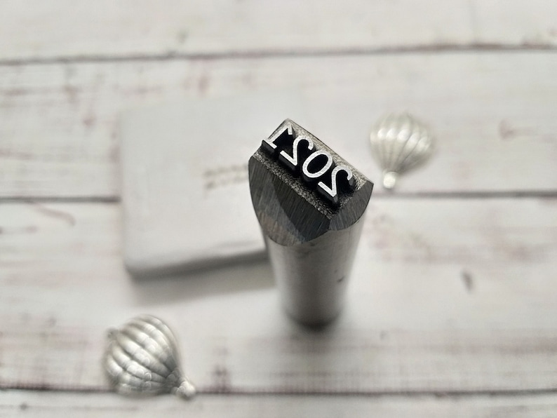 May include: A close-up of a metal stamp with the year 2021 engraved on it. The stamp is silver and has a rounded top.