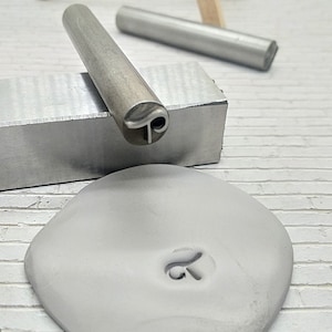 May include: A metal stamp with a swirl design is used to create an impression in a piece of gray clay. The stamp is resting on a metal block.