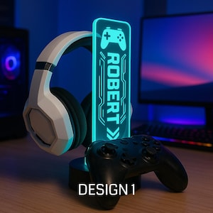 May include: A gaming headset stand with a personalized LED light. The light is teal and features a controller icon and the name "ROBERT". A black game controller and white headset are also present. The base of the stand is black, and the design is labeled "DESIGN 1".